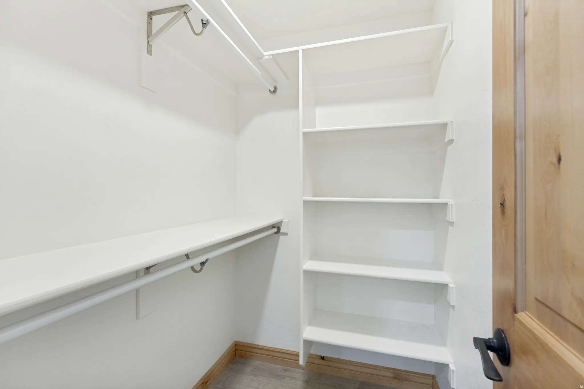 Walk-in closet featuring built-in shelving, white walls, wood-finish flooring, and a natural wood door with a matte black handle