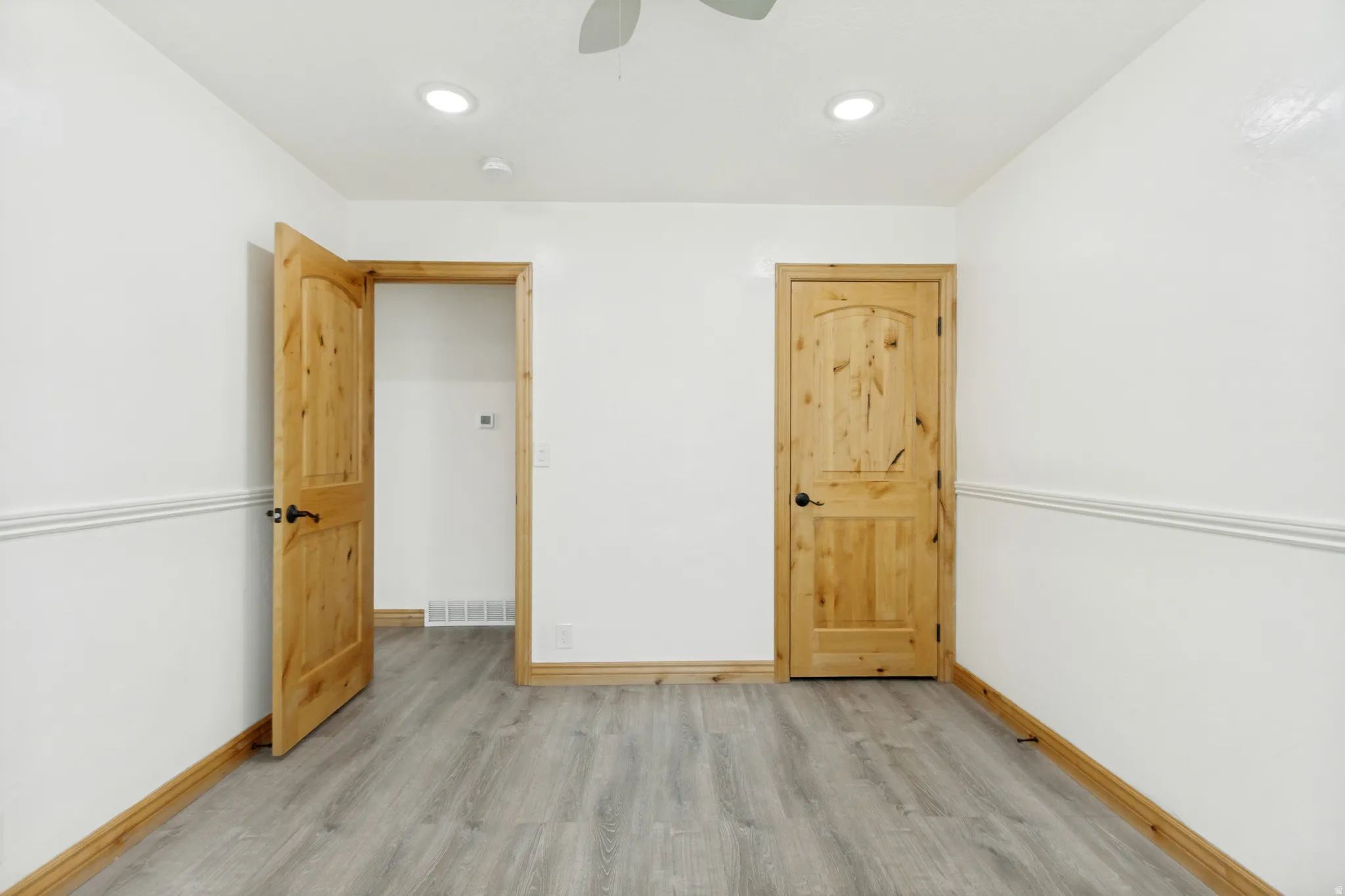 Interior room featuring two wooden doors with arched top panels, wood-finish flooring, white walls with chair rail molding, and recessed ceiling lighting
