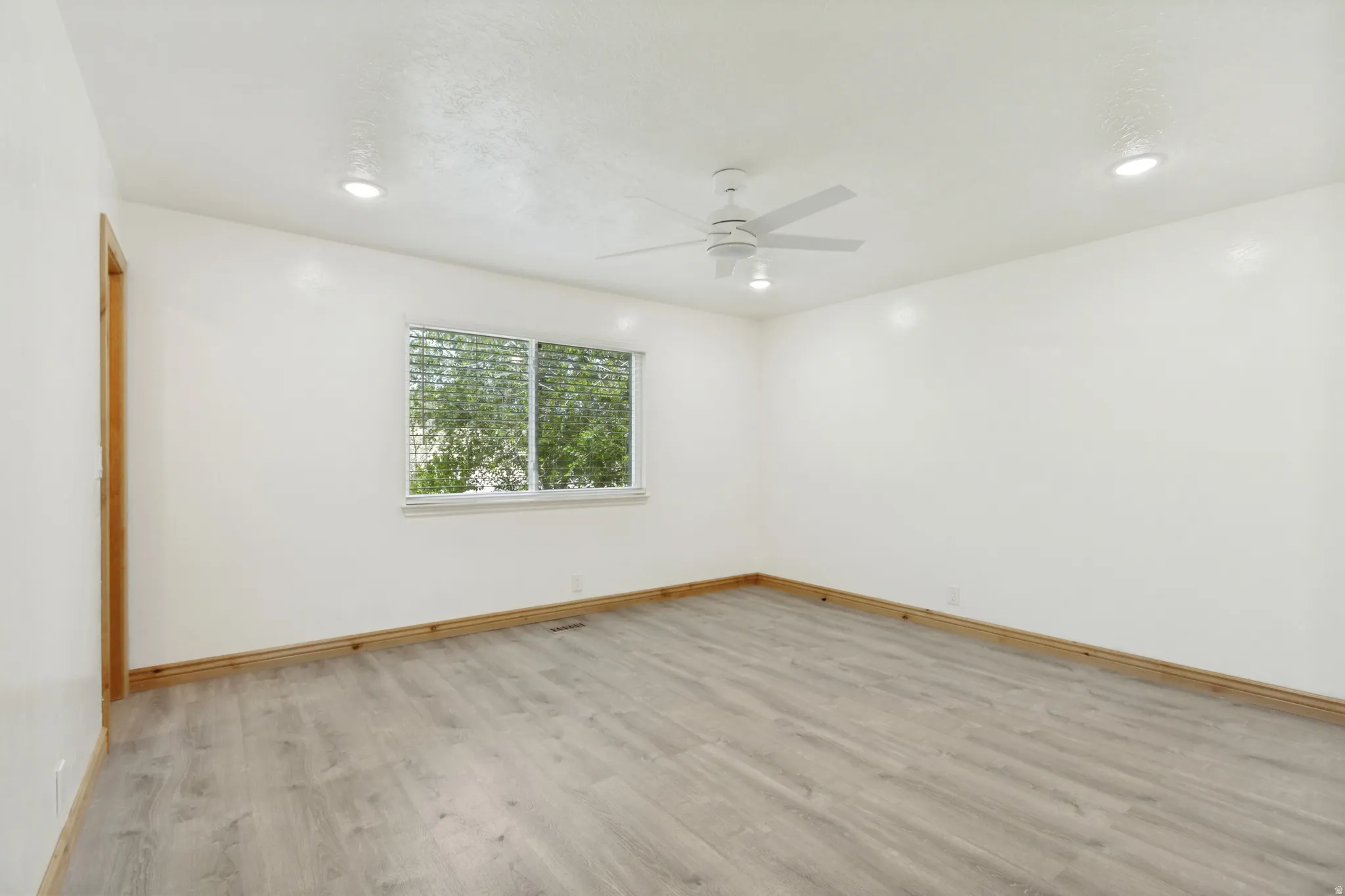Spacious room featuring light wood-finish flooring, a white ceiling fan, recessed lighting, a window with horizontal blinds, and natural wood trim