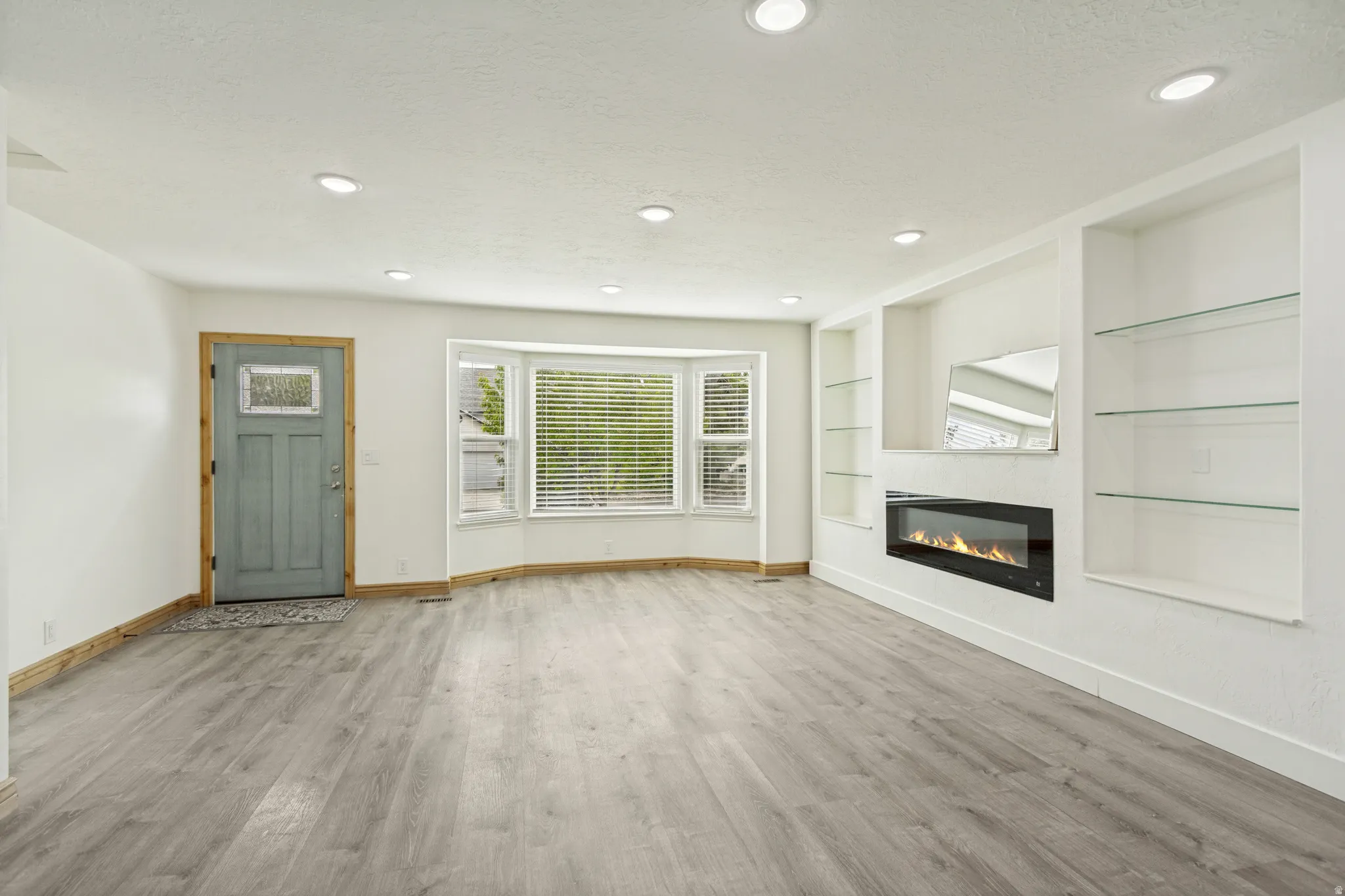 Spacious living area featuring light wood-finish flooring, a contemporary electric fireplace with built-in shelving, a bay window with blinds, recessed ceiling lighting, and a paneled entry door