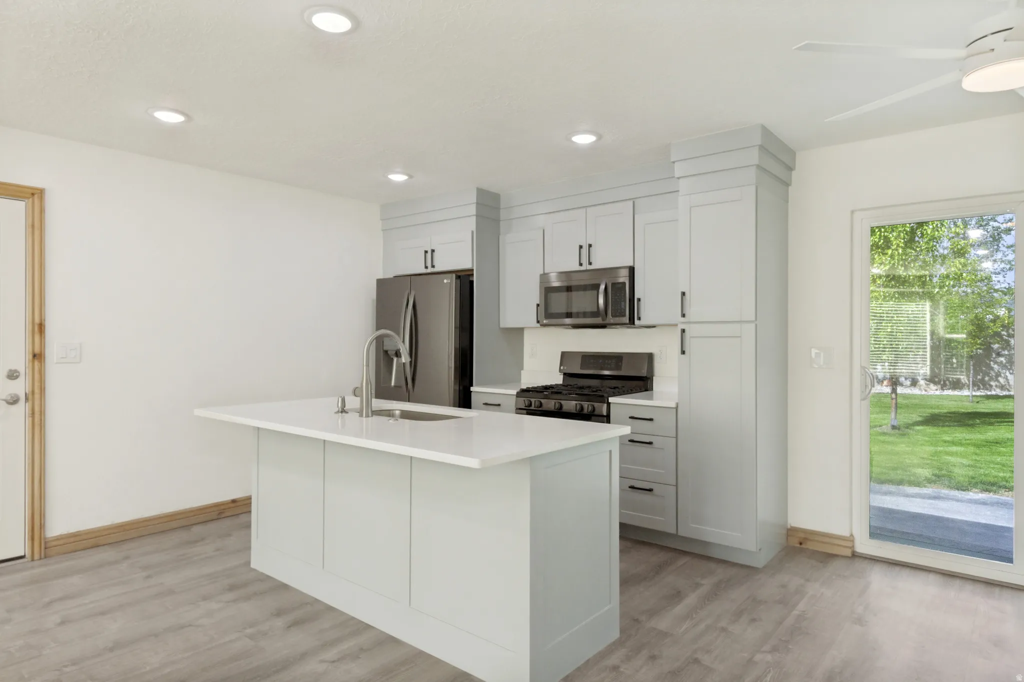 Bright kitchen featuring a central island with a sink, light gray cabinetry, stainless steel appliances, and wood-finish flooring