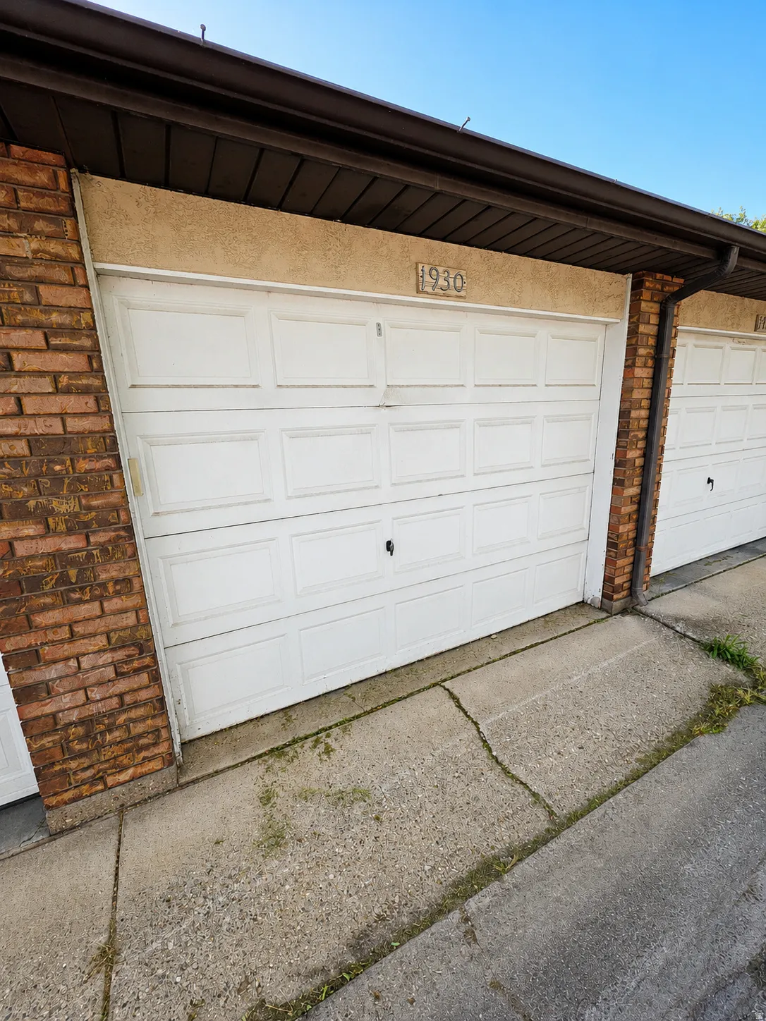 White paneled automatic  garage door with an overhead brown gutter system, textured stucco facade, and brick pillar detailing