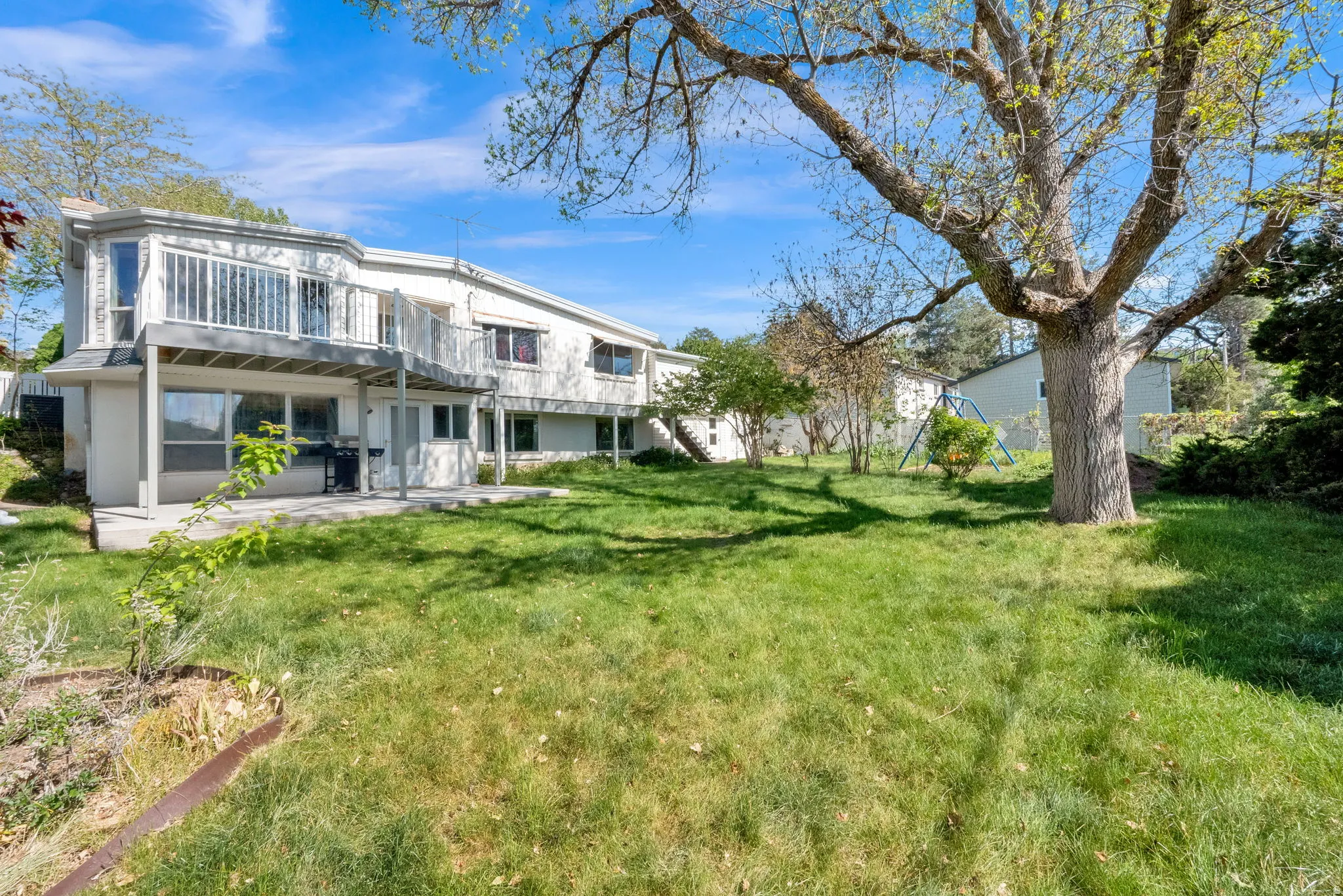 Expansive rear yard featuring a multi-level white exterior, upper-level balcony with white railings, lower-level patio, and a mature tree