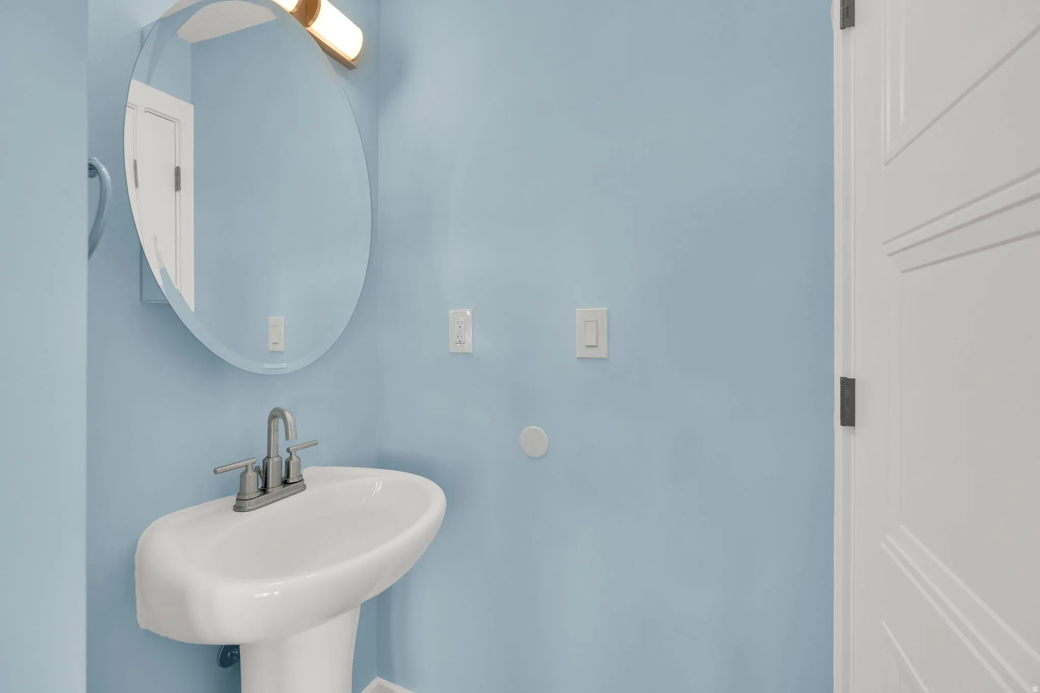 Pedestal sink with a two-handle faucet, oval mirror, and contemporary vanity lighting