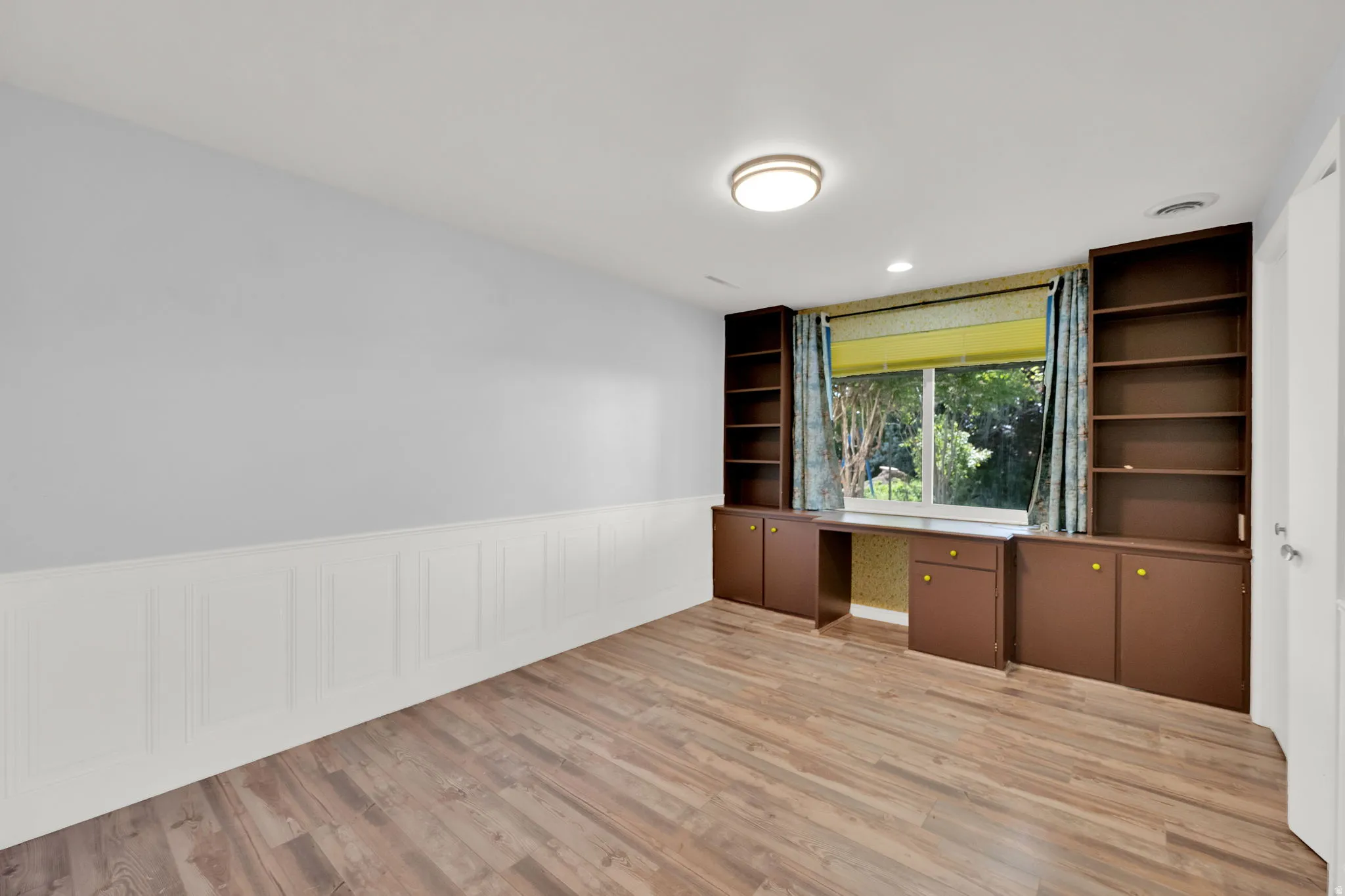 Room featuring wood-finish flooring, wainscoting, and a window with a green outdoor backdrop