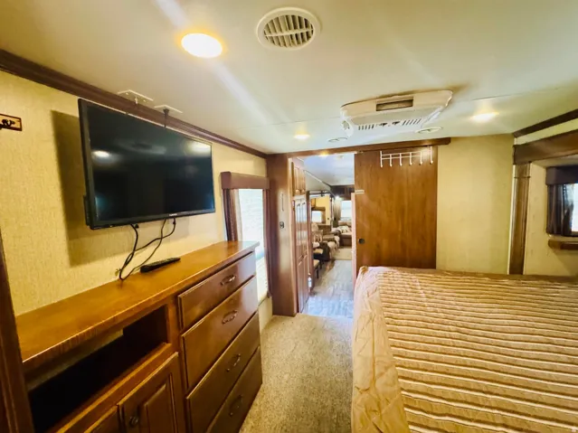 Bedroom featuring a mounted flat-screen television, a built-in dresser with drawers and open shelving, a sliding wood-finish door, overhead recessed lighting, and a ceiling-mounted air conditioning unit