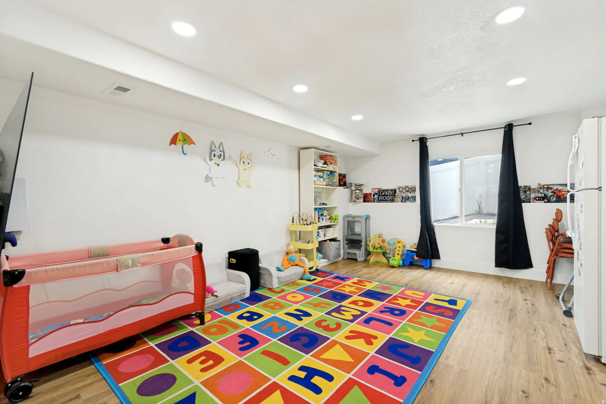 Versatile lower-level room featuring wood-finish flooring, recessed lighting, a built-in shelving unit, and a window with black privacy drapes