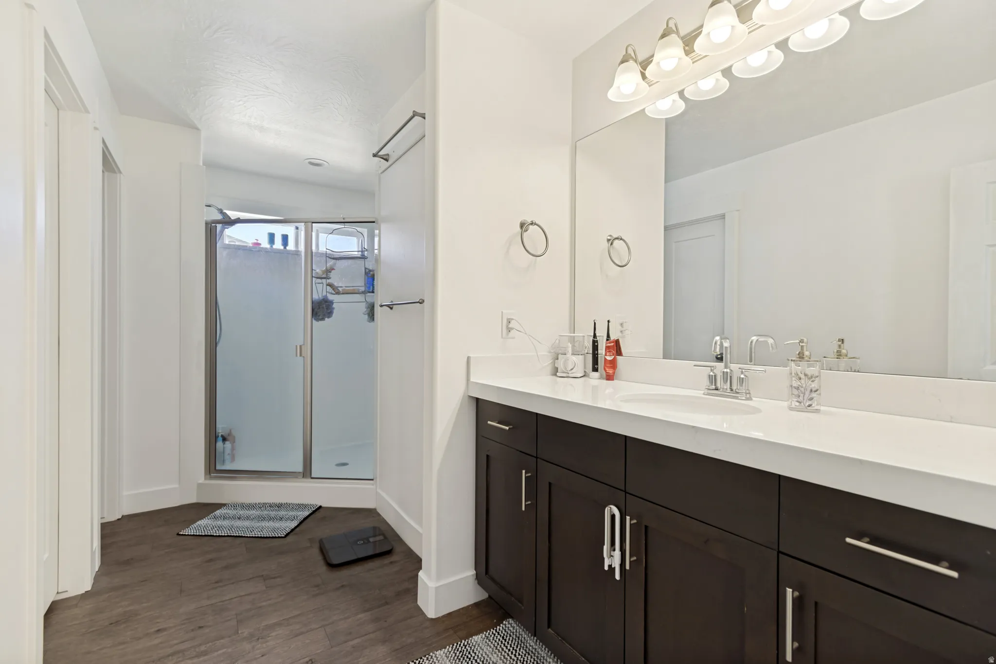 Bathroom featuring a wood-finish floor, a shower with a sliding glass door, a double vanity with dark wood cabinetry, a white countertop, and a large mirror with overhead lighting