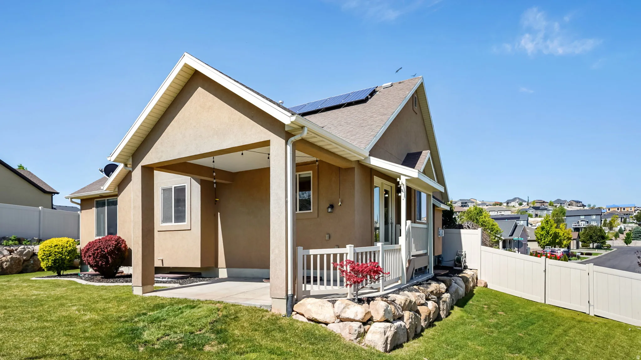 Rear exterior featuring a covered patio, solar panels, a tiered landscape with natural rock hardscaping, and an additional elevated patio with white railing