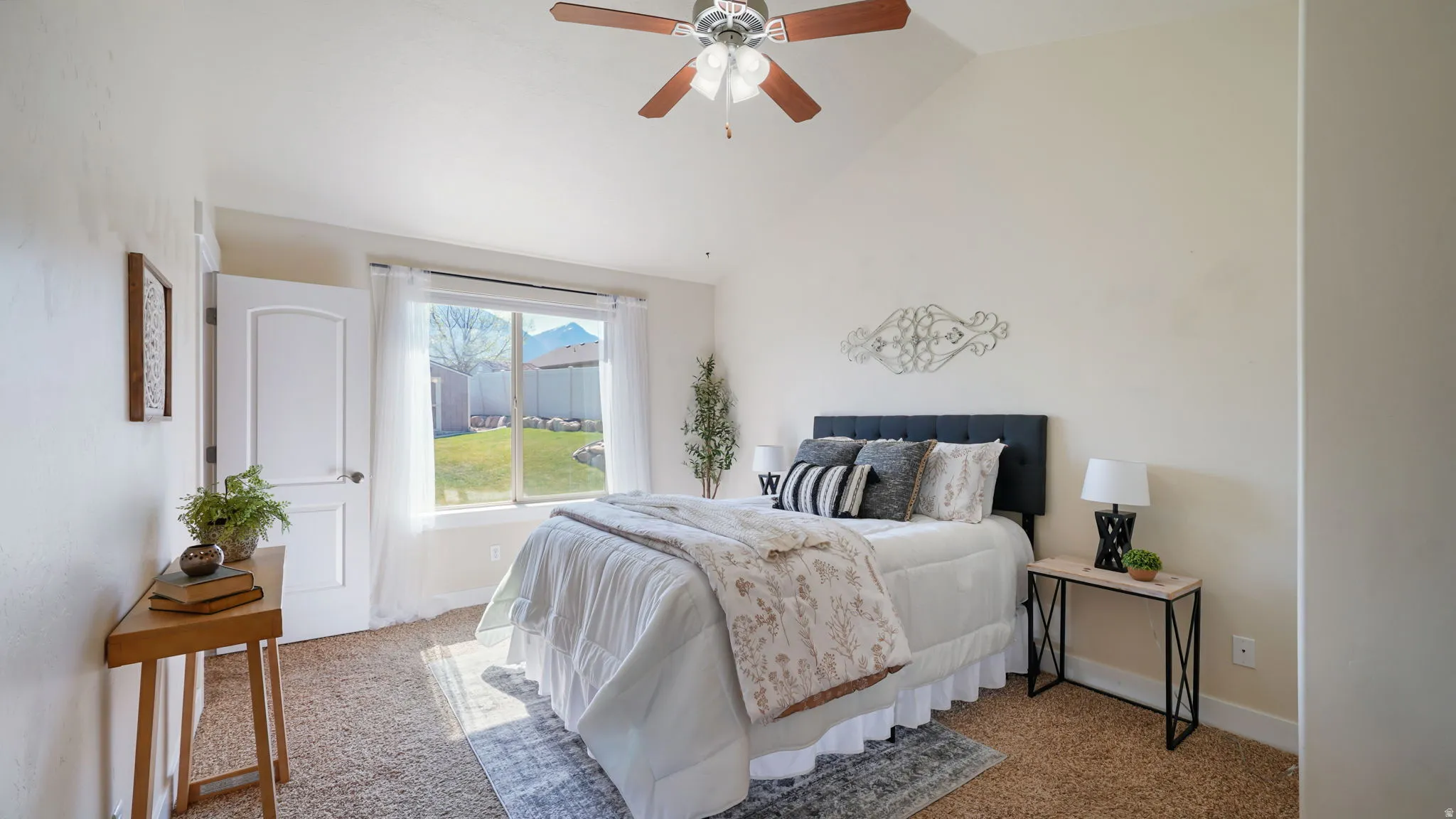 Carpeted bedroom featuring a vaulted ceiling with a ceiling fan, a window overlooking a fenced yard, and a single pane door
