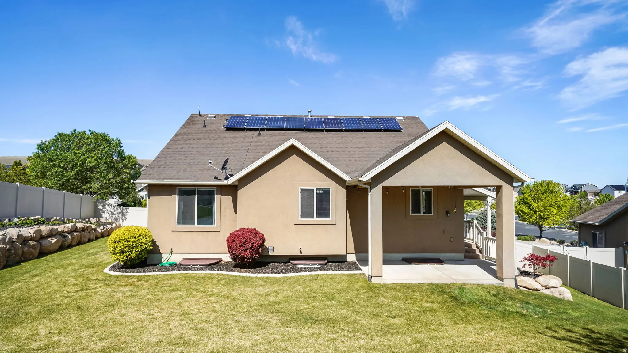 Rear exterior featuring a covered patio, solar panels, a rock retaining wall, and a manicured lawn