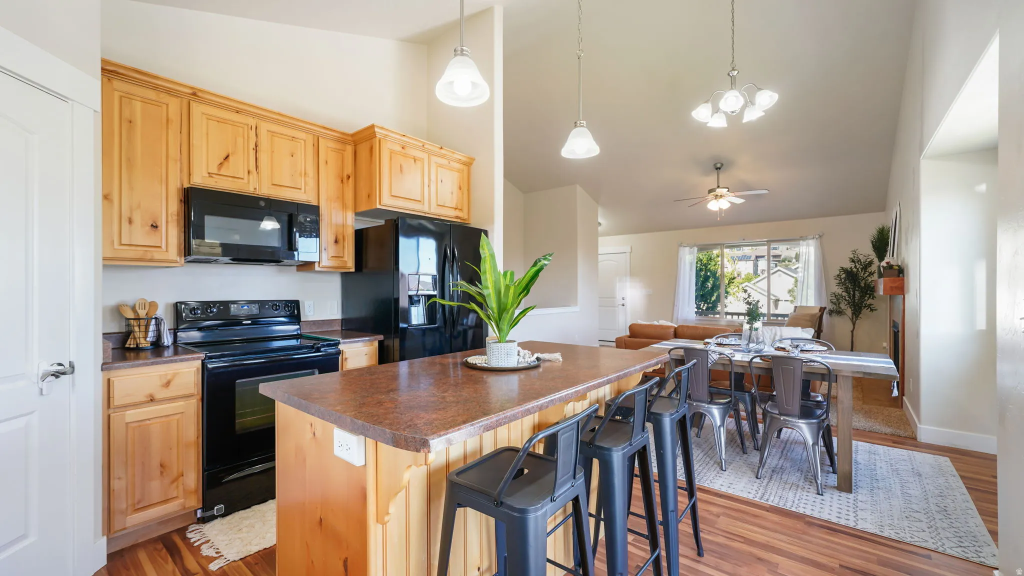 Open-concept kitchen featuring light wood cabinetry, black appliances, a central island with bar seating, and wood-finish flooring