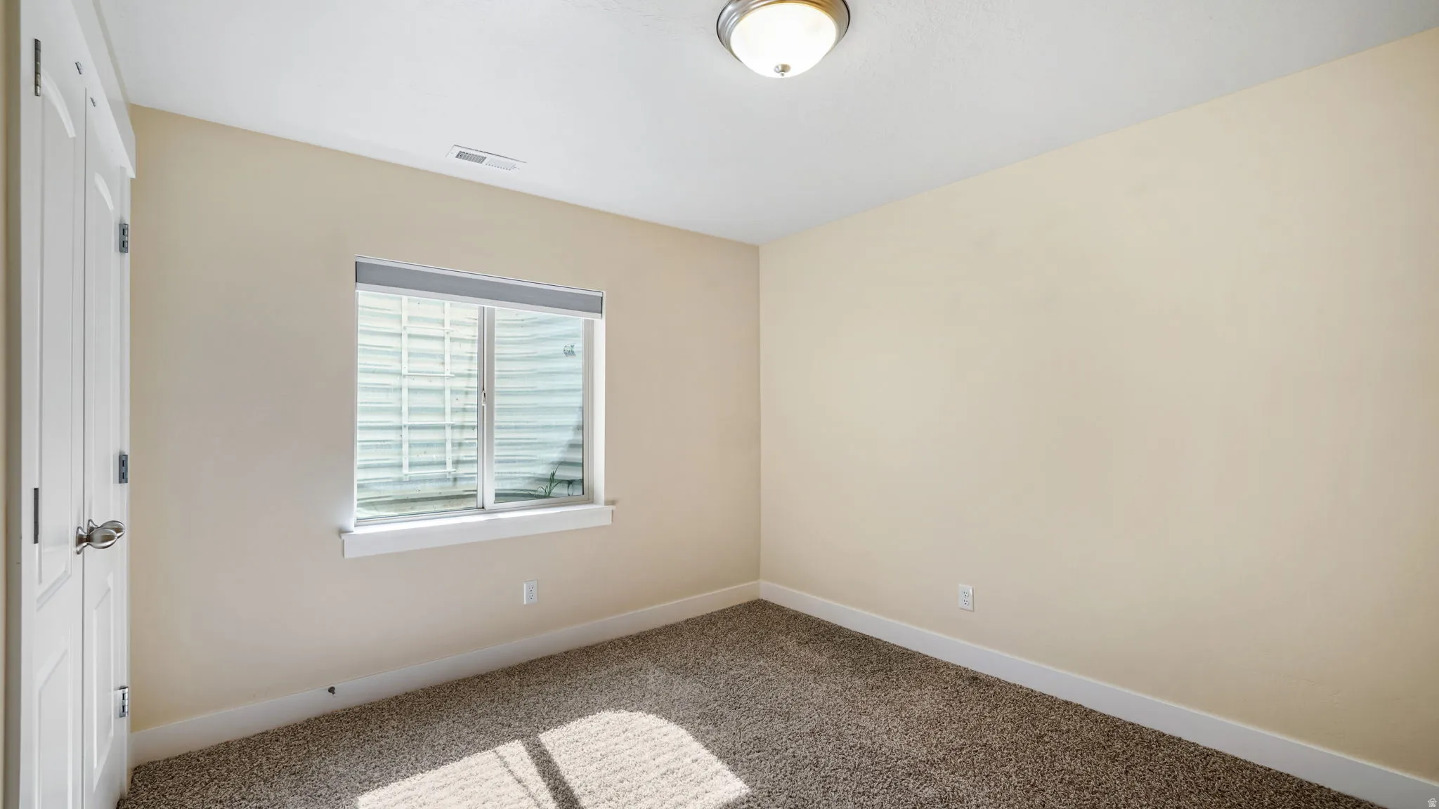 Inviting room featuring neutral tone walls, patterned carpet flooring, a single window with a window well, white trim, and a flush mount ceiling light