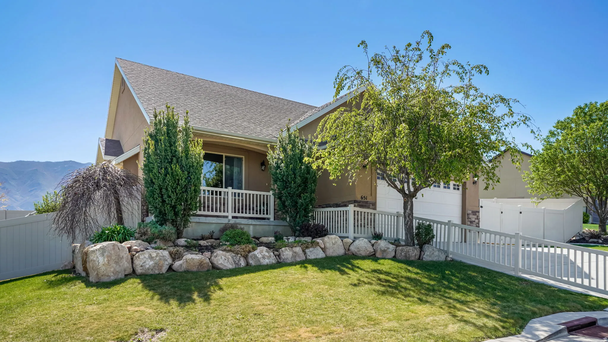 Stucco exterior residence with a composition shingle roof, featuring a front porch with white railing, a two-car garage, and a rock garden with mature landscaping