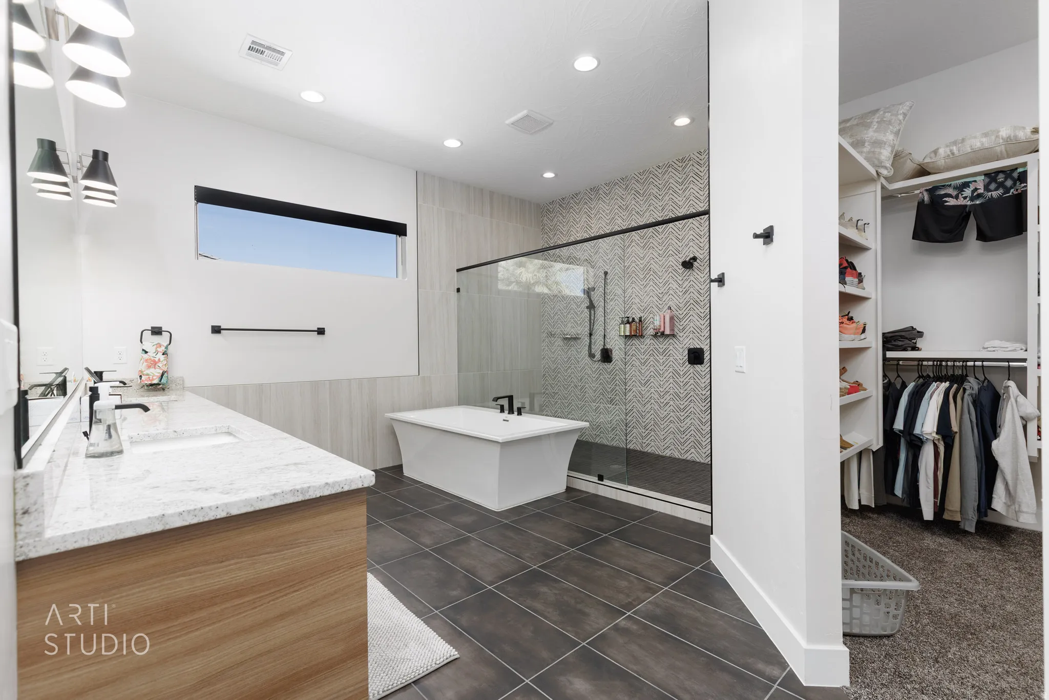 Contemporary bathroom featuring a freestanding tub, glass-enclosed shower with chevron tile, wood-finish vanity with dual sinks, and a walk-in closet with built-in shelving