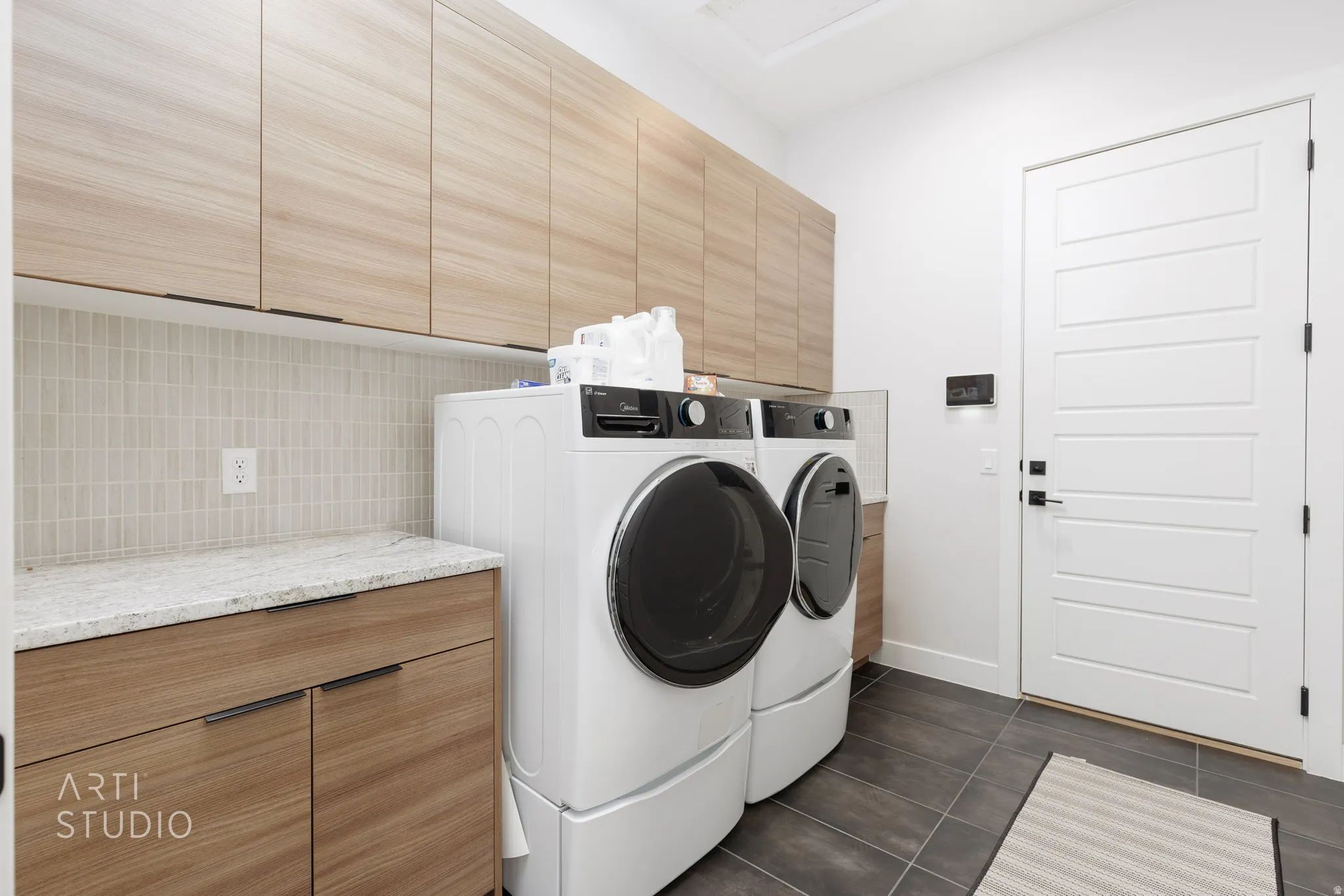 Dedicated laundry space featuring extensive upper and lower wood-finish cabinetry, a light-toned mosaic tile backsplash, a stone countertop, and dark-toned floor tiling