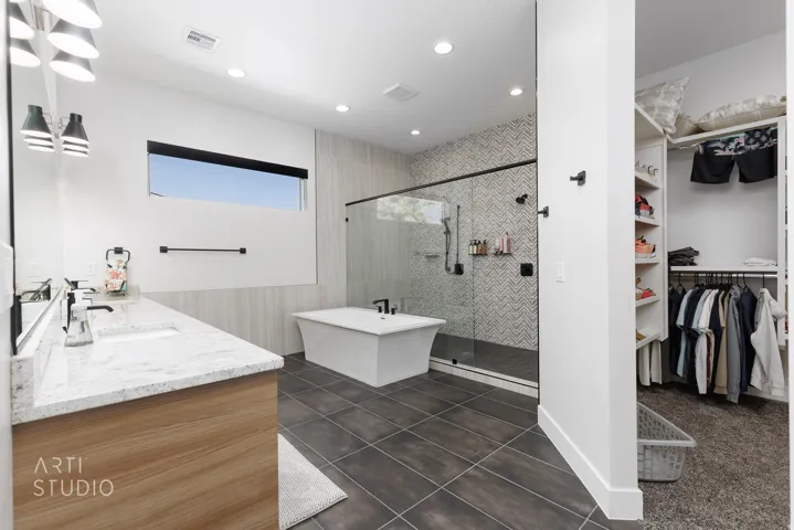 Contemporary bathroom featuring a freestanding tub, glass-enclosed shower with chevron tile, wood-finish vanity with dual sinks, and a walk-in closet with built-in shelving