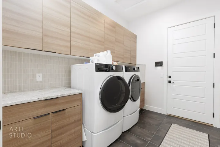 Dedicated laundry space featuring extensive upper and lower wood-finish cabinetry, a light-toned mosaic tile backsplash, a stone countertop, and dark-toned floor tiling