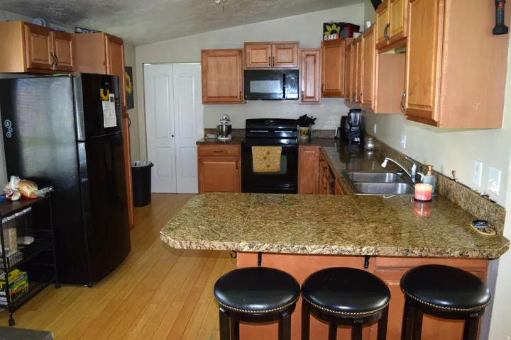 Kitchen featuring wood-finish flooring, a vaulted ceiling, wood cabinetry, dark-toned appliances, and granite-style countertops with an integrated breakfast bar