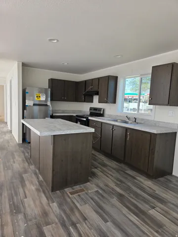 Contemporary kitchen featuring dark wood-finish cabinetry, a central island with a light-toned countertop, stainless steel appliances, recessed ceiling lighting, and wood-finish flooring