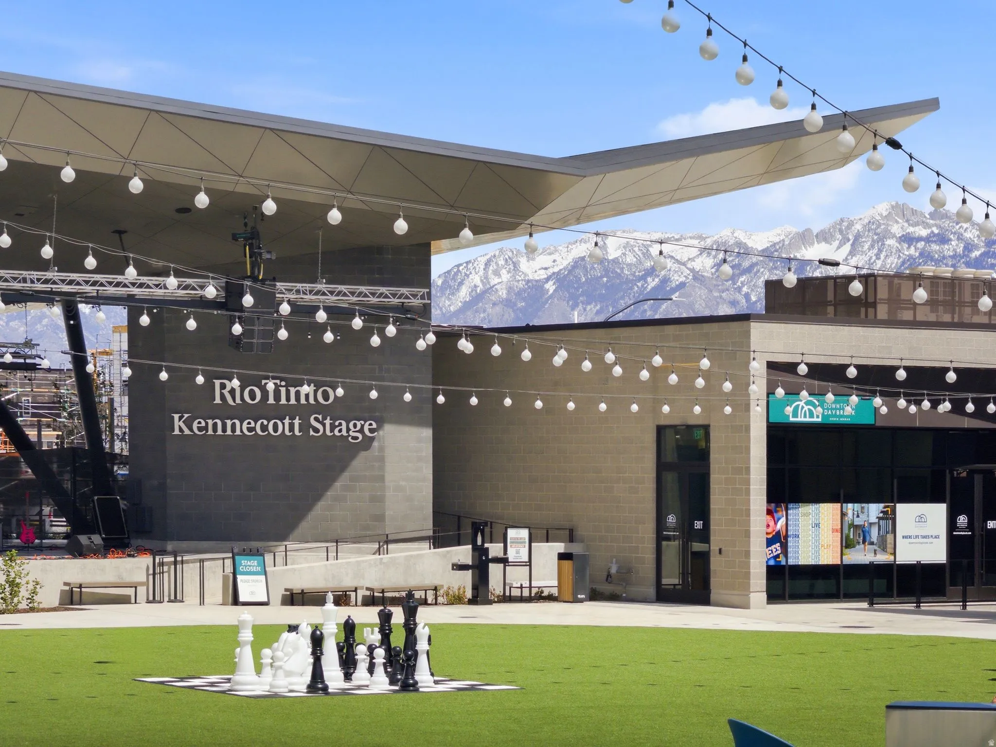 Outdoor entertainment area featuring a large stage with a modern canopy, string lighting overhead, a turf lawn, and oversized chess pieces