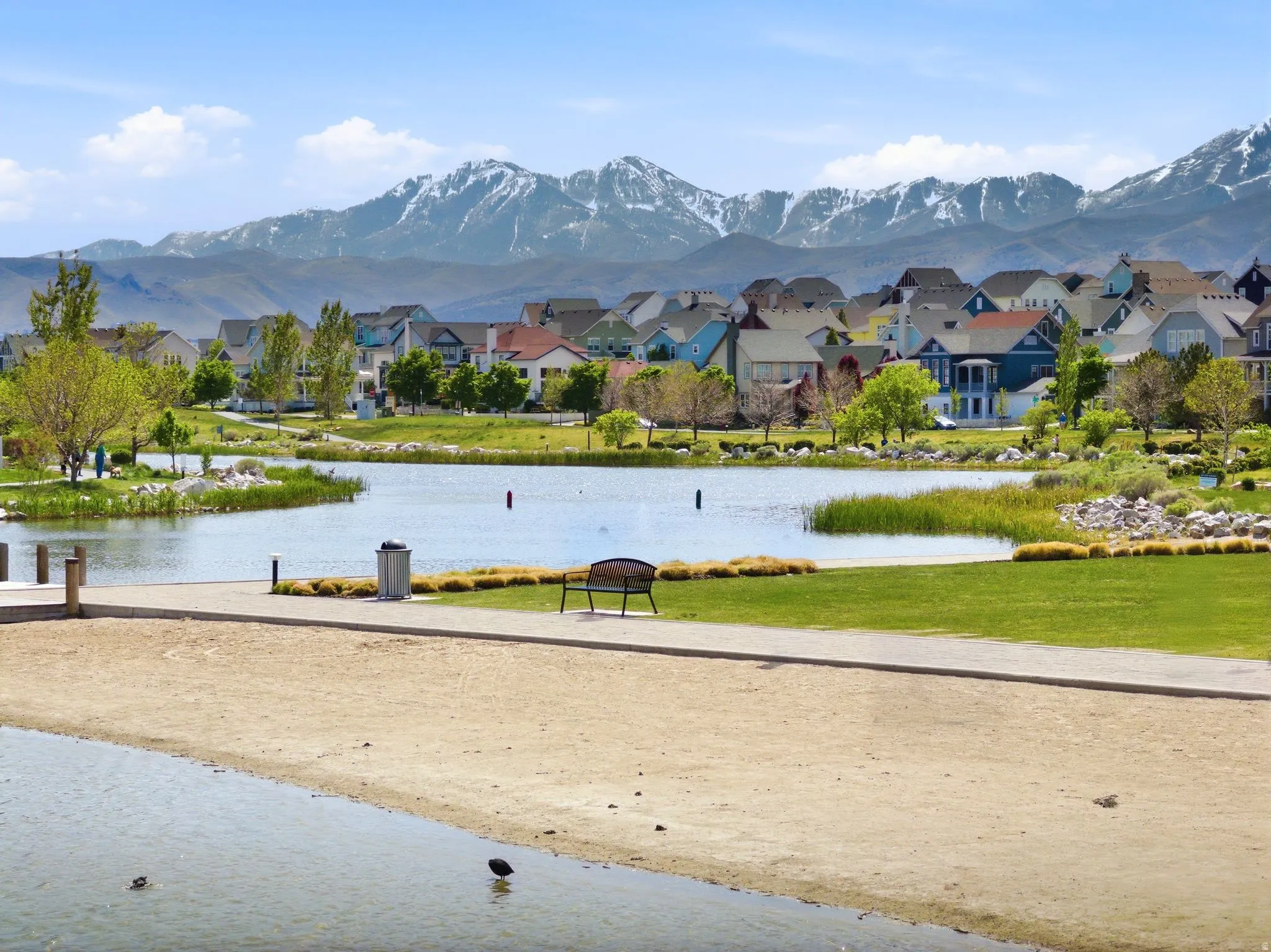 Waterfront property featuring a sandy shore, manicureod green space, and a community of homes against a mountain backdrop with trails to use to enjoy.