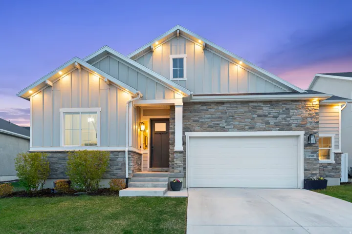 Contemporary home exterior featuring board and batten siding, stone accents, a two-car garage, an illuminated entryway, and a manicured lawn