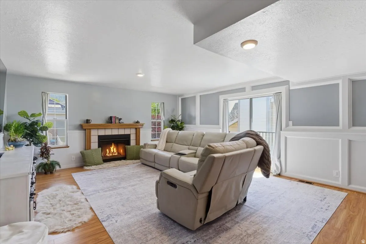 Living area featuring a central fireplace with a wood mantle and tile surround, wood-finish flooring, two large windows, sliding glass door access, and decorative wall paneling
