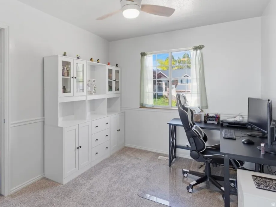 White-painted room extending from primary can be used as closet or office, even a library! featuring wainscoting,  a window with exterior views, a ceiling fan with integrated lighting, and carpet flooring