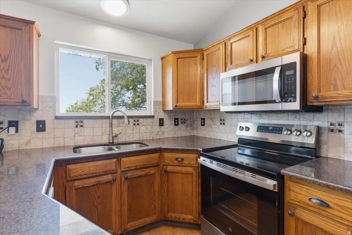 Kitchen featuring wood cabinetry, stainless steel appliances, a double basin sink, stone countertops, and a tiled backsplash