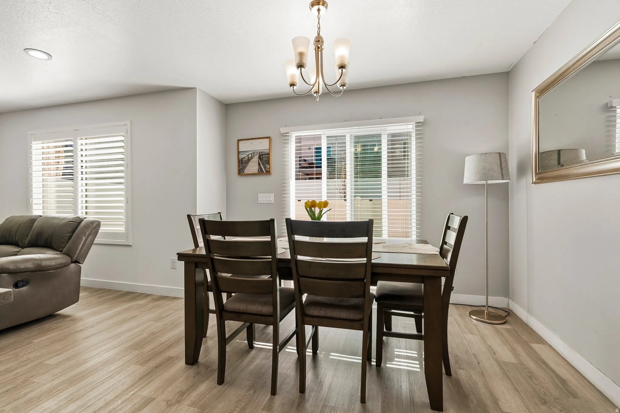 Dining area with wood-finish flooring, two window openings with horizontal blinds, and a brass-tone chandelier