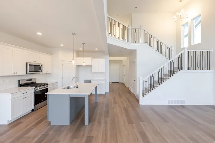 Open-concept living area featuring a kitchen with white cabinetry, stainless steel appliances, a center island with sink, wood-finish flooring, and a grand staircase with decorative railing