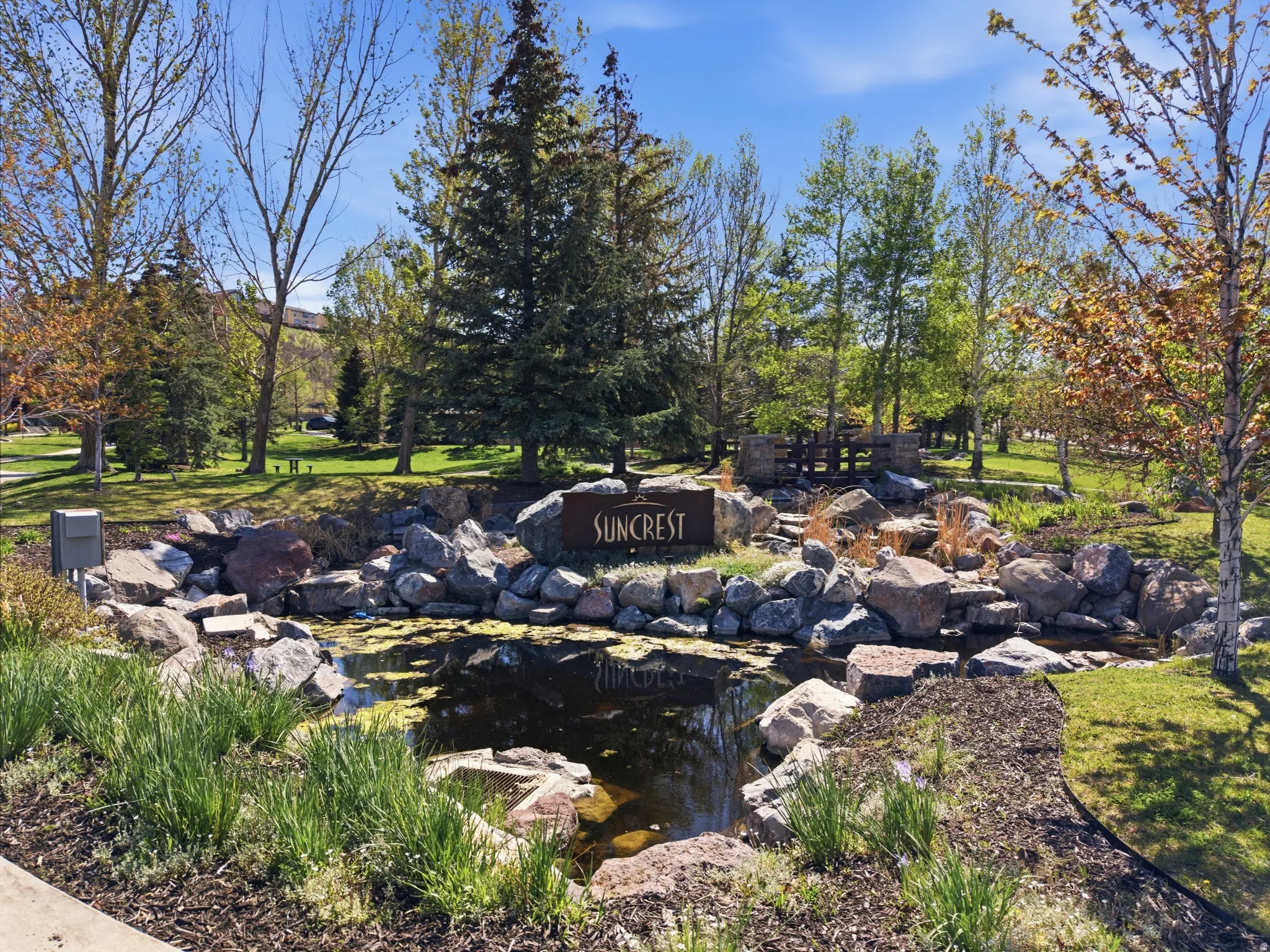 Community entrance featuring a water feature, stone landscaping, mature trees, a footbridge, and a "Suncrest" monument