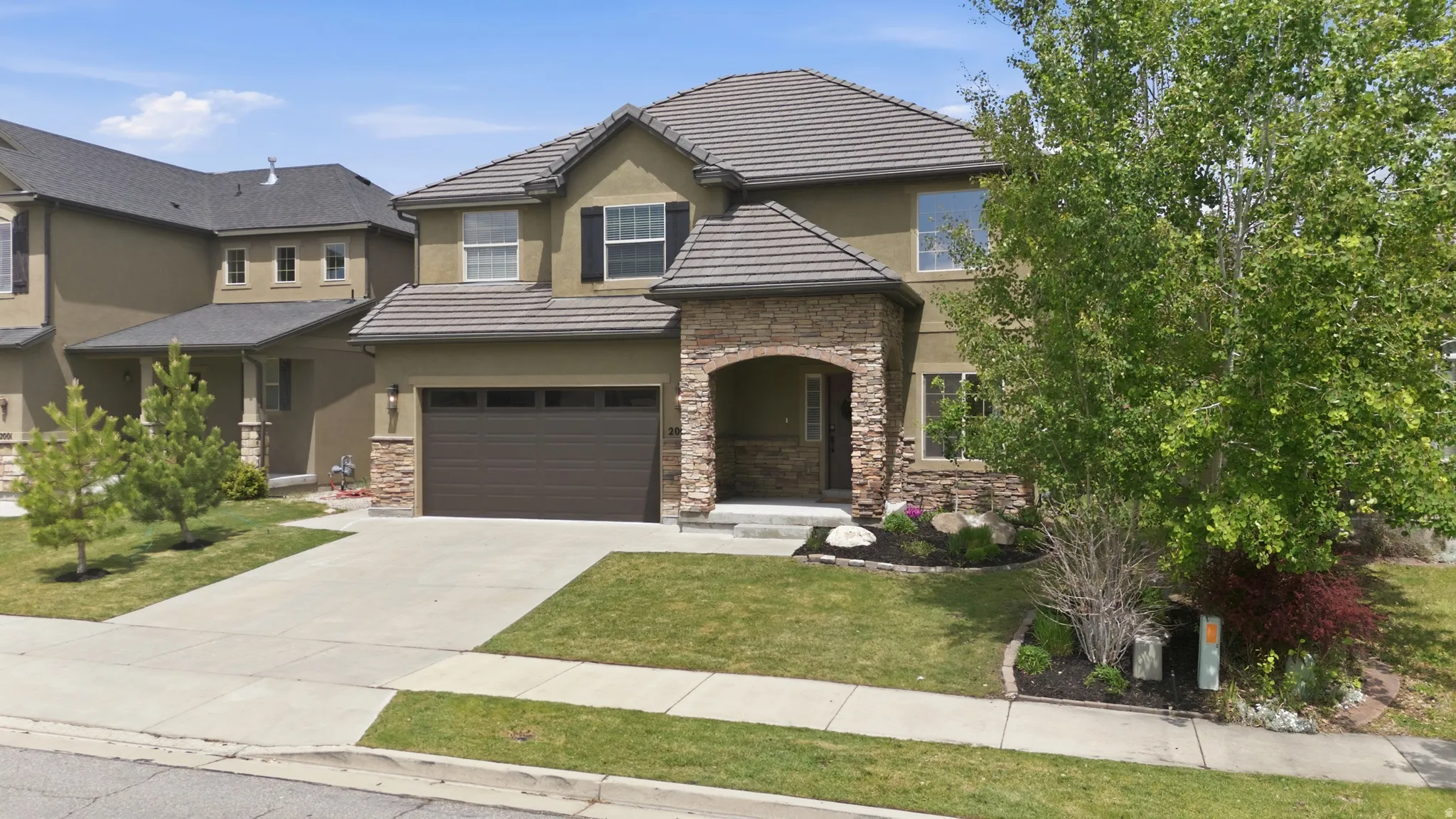 Two-story residence featuring a stucco exterior with stone accents, a tile roof, an attached two-car garage, mature landscaping, and a concrete driveway