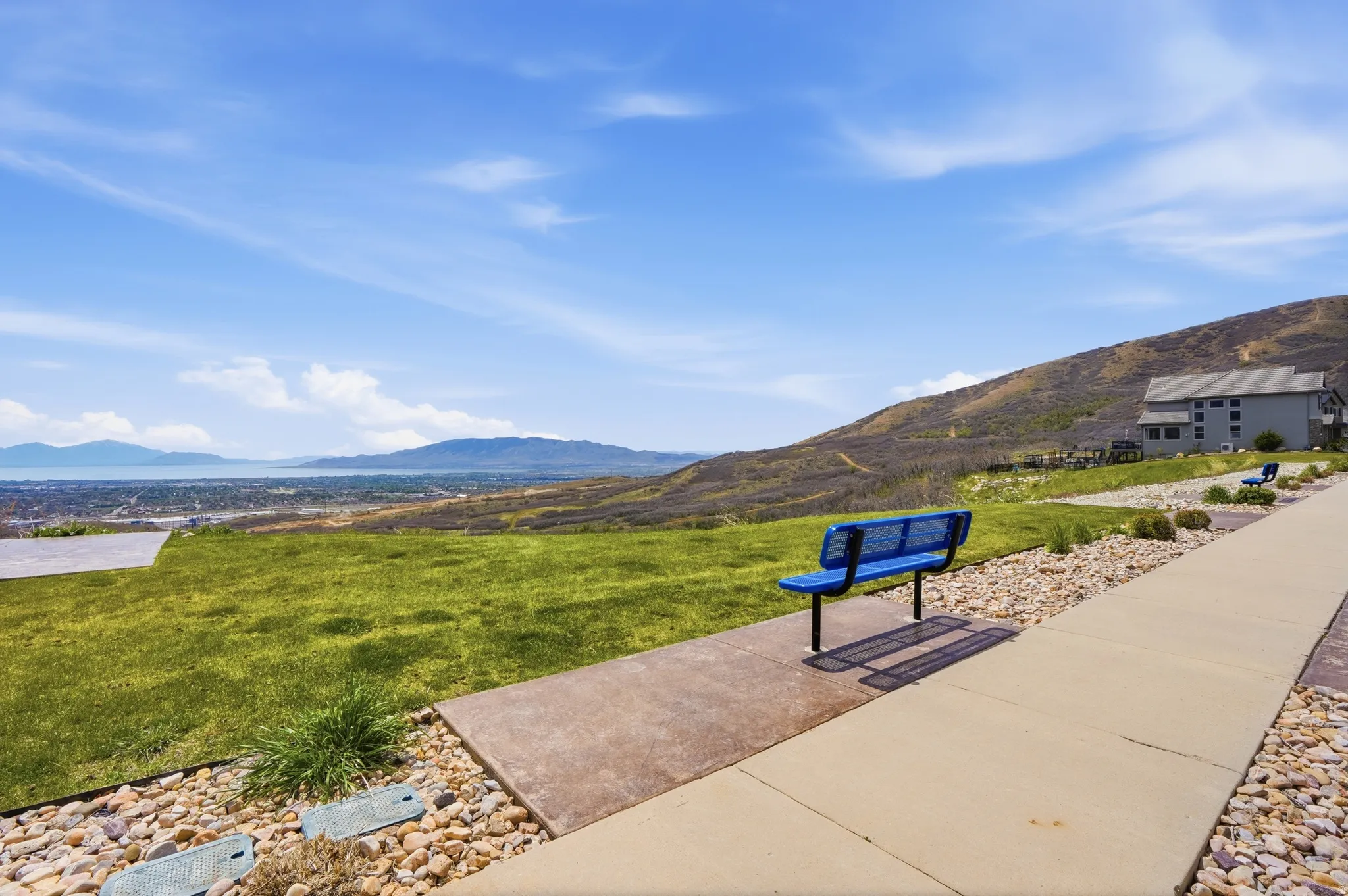 Expansive green space featuring a concrete pathway, blue metal bench, decorative rock landscaping, and distant mountain and water scenery
