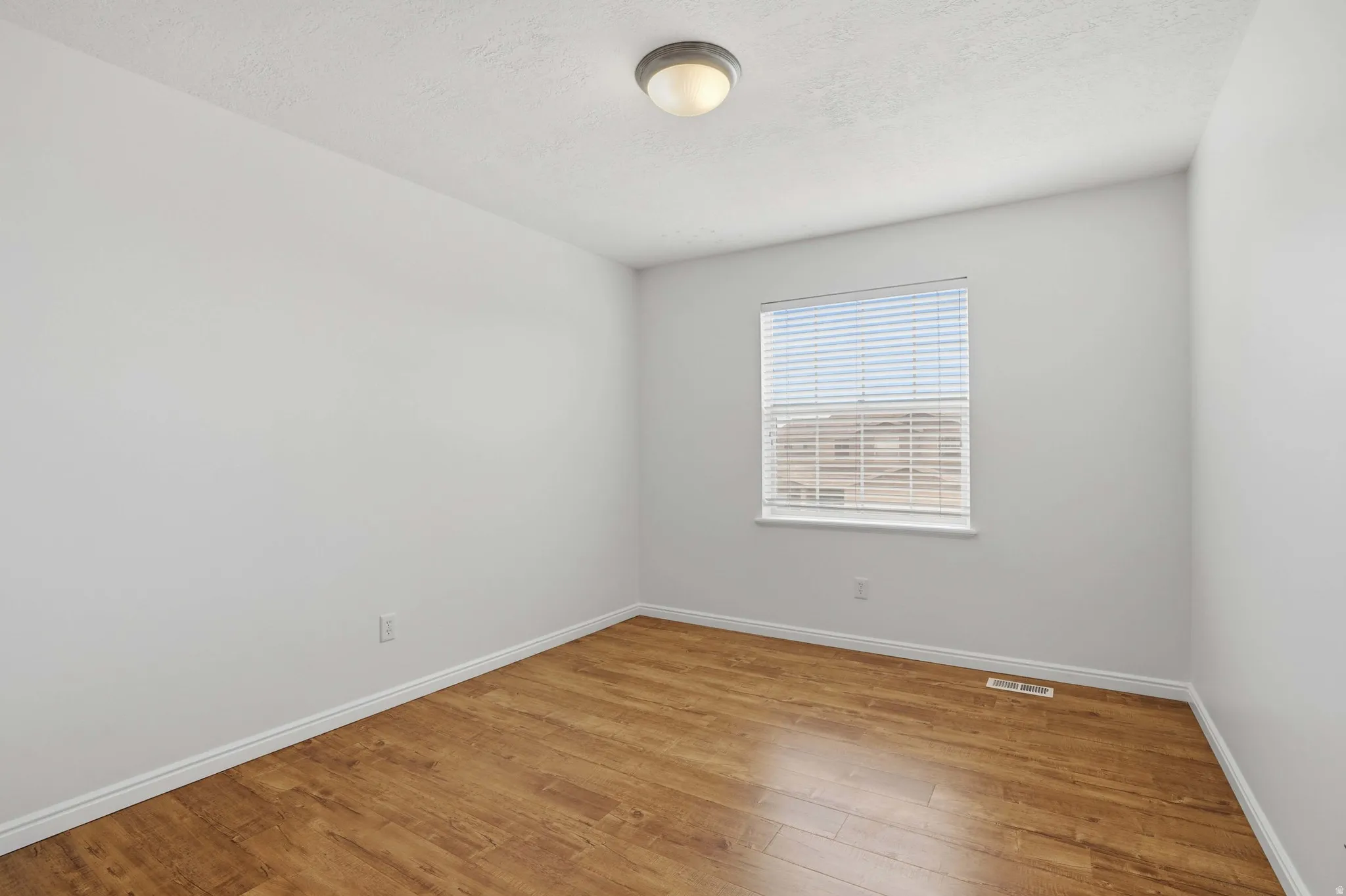 Neutral interior with wood-finish flooring, white baseboards, a single window with blinds, and a ceiling-mounted light fixture