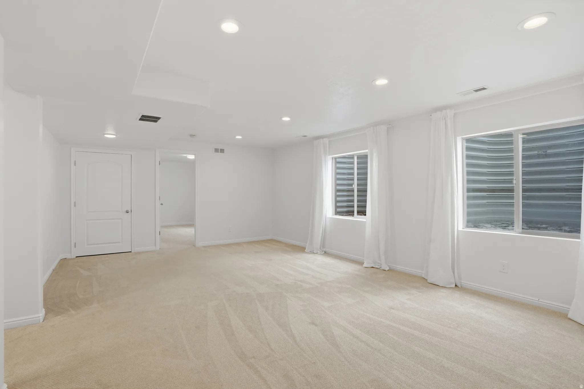 Spacious room featuring neutral carpeting, recessed ceiling lighting, two egress windows with white drapes, and white trim throughout