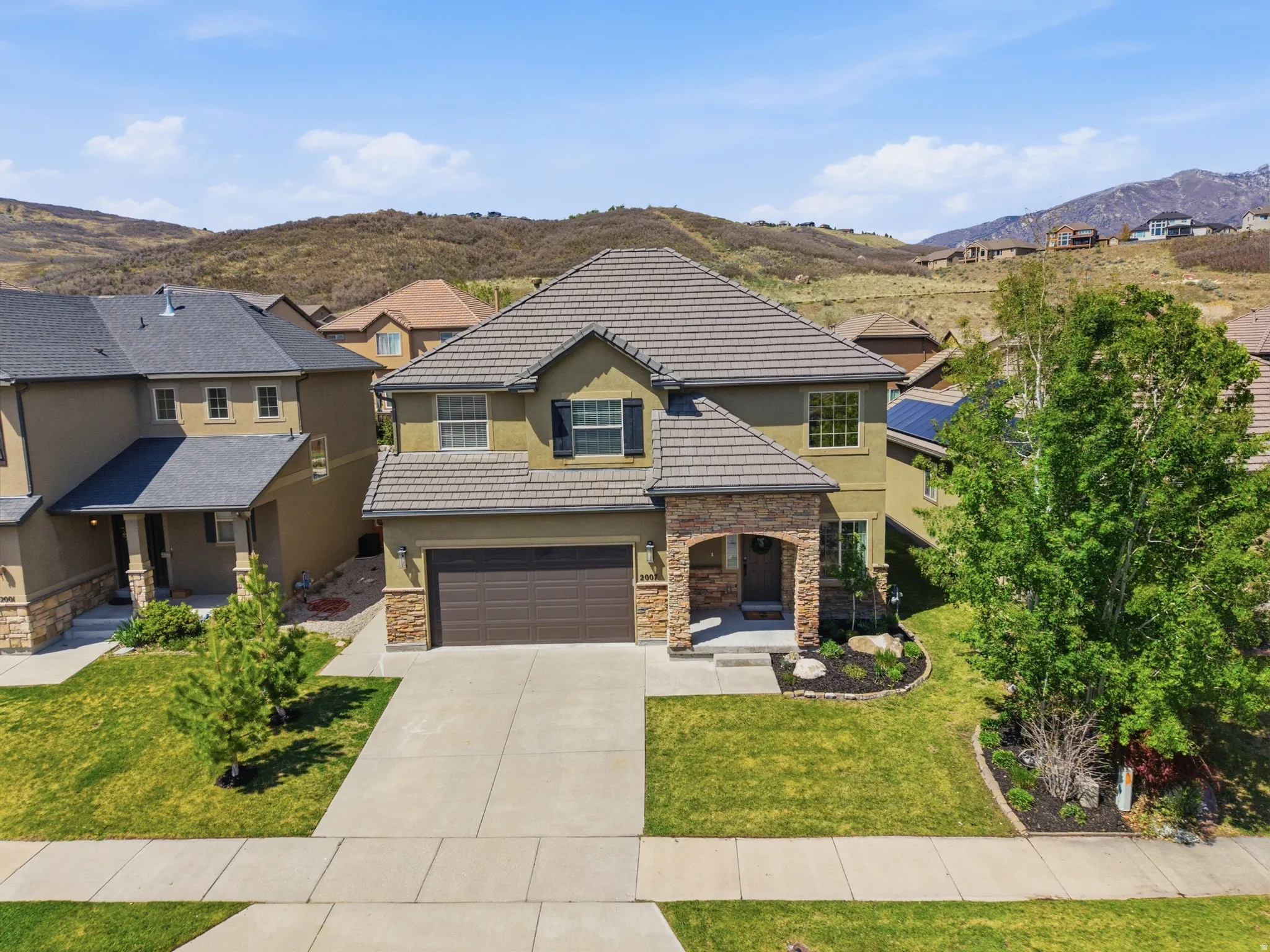 Two-story residence featuring a tile roof, stone and stucco exterior, an integrated two-car garage, and landscaped front yard with mature trees