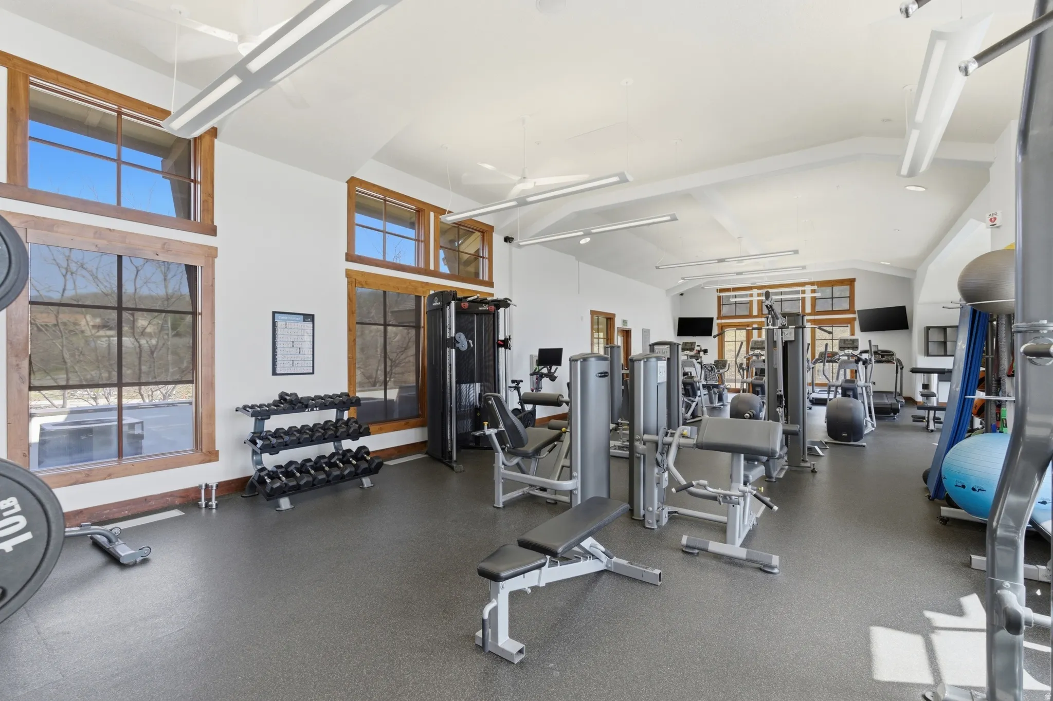 Spacious fitness center featuring high vaulted ceilings, expansive windows with wood trim, resilient rubber flooring, and integrated overhead lighting
