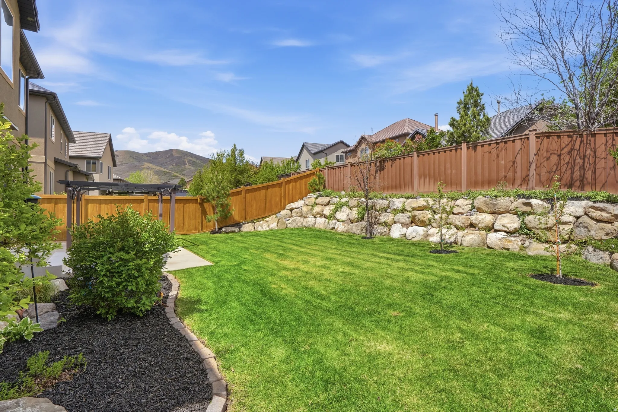 Expansive backyard featuring a lush lawn, a tiered retaining wall with natural stone, a full privacy fence, a concrete patio, and a dark wood-finish pergola