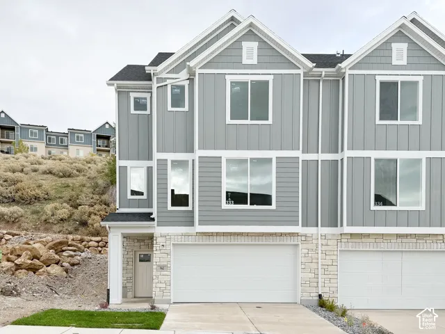 Multi-story townhouse exterior featuring gray vertical and horizontal siding, a stone-veneer foundation, a dual-gabled roofline, and an attached two-car garage
