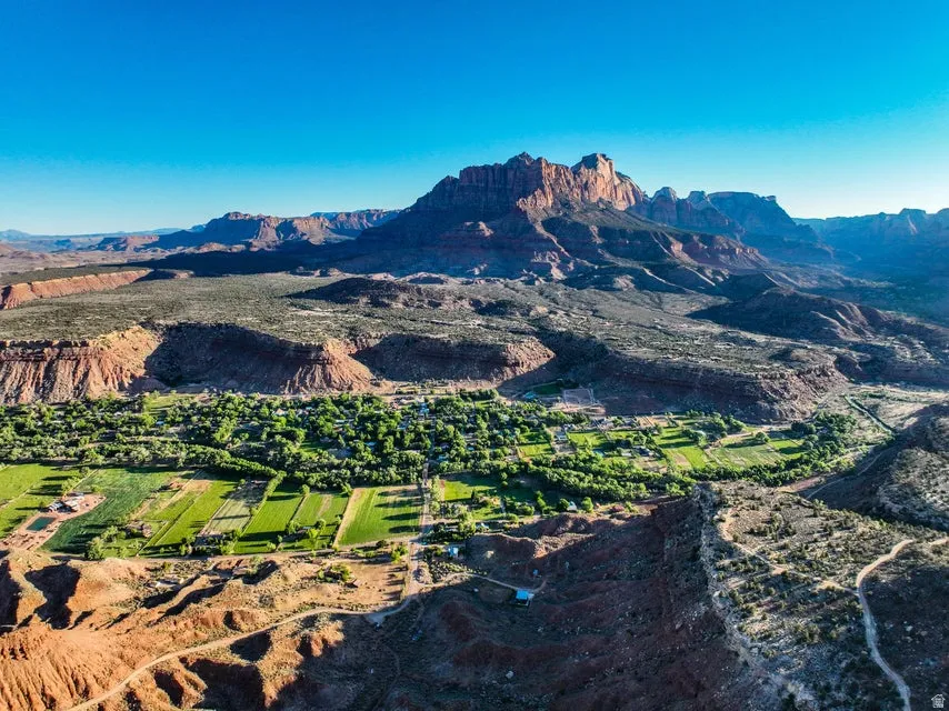 Expansive aerial landscape featuring a vibrant green valley with agricultural fields and clustered structures, set against a backdrop of towering red rock formations and a clear blue sky