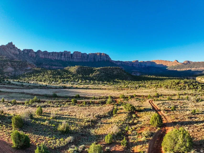 Expansive desert landscape featuring rugged mountain formations, a broad valley floor, scattered green shrubs, and a distinct dirt path