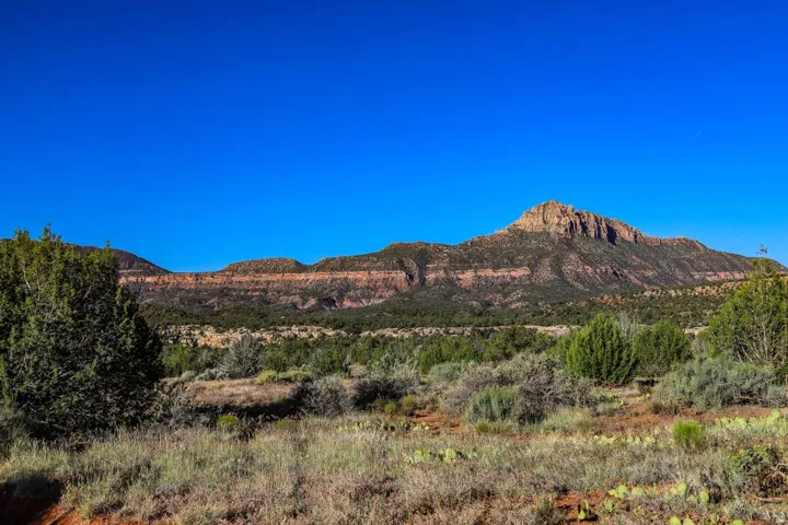 Expansive natural landscape featuring a prominent mountain formation, varied low-lying vegetation, a clear blue sky, and distant rock formations