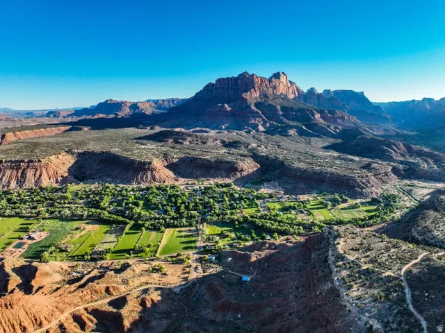 Expansive aerial landscape featuring a vibrant green valley with agricultural fields and clustered structures, set against a backdrop of towering red rock formations and a clear blue sky