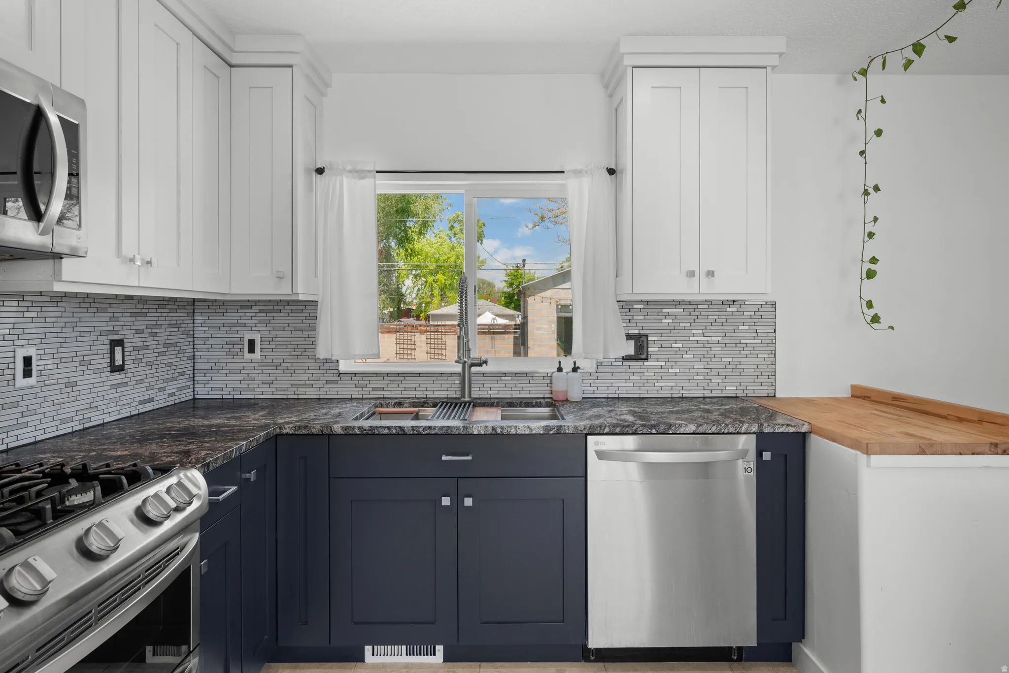 Kitchen featuring two-tone cabinetry with white upper and dark lower units, stainless steel appliances, a mosaic tile backsplash, a dark stone-finish countertop, and a wood-finish peninsula surface
