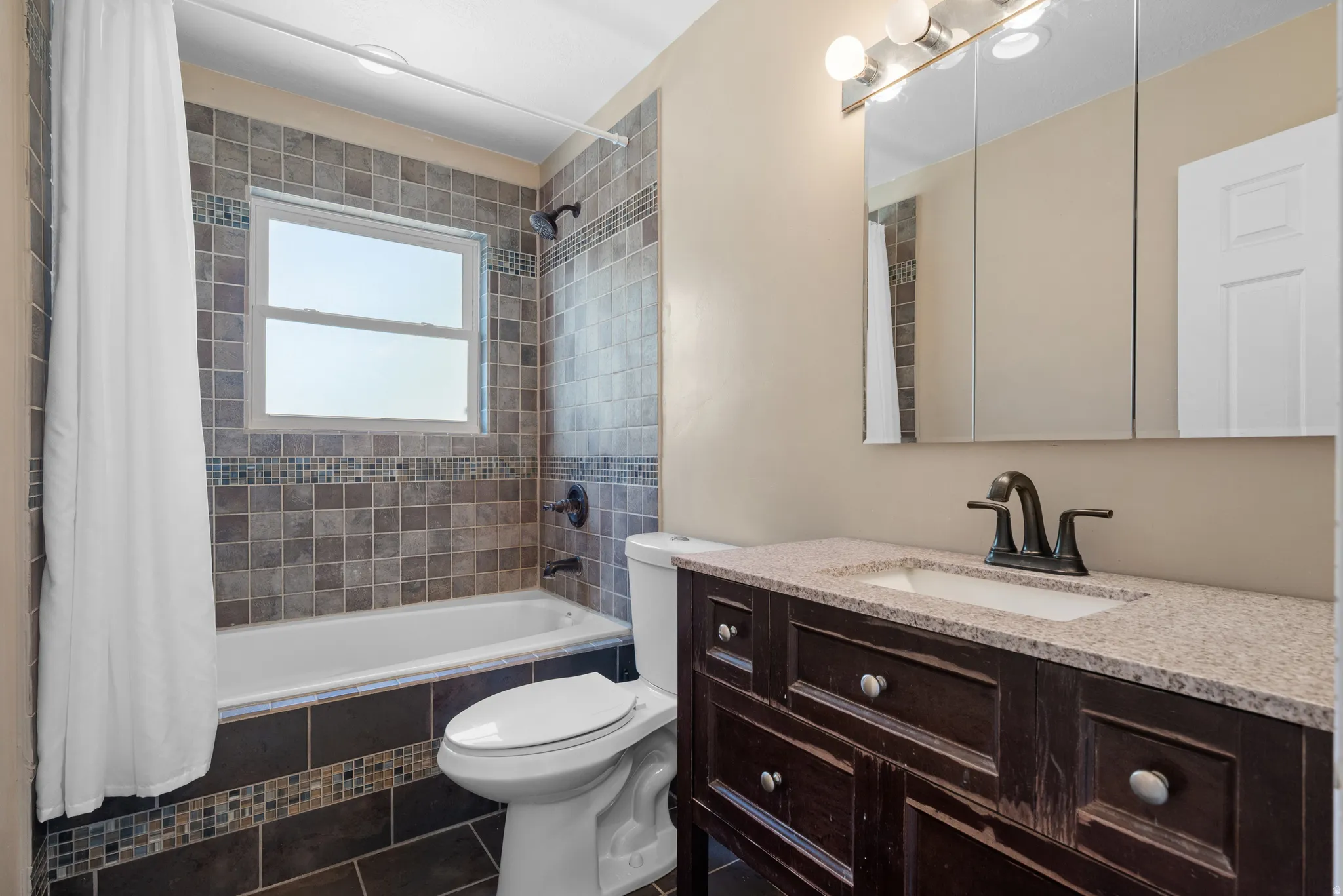 Full bathroom featuring a window, tile shower surround, dark wood-finish vanity with a light-toned countertop, and bronze-finish fixtures