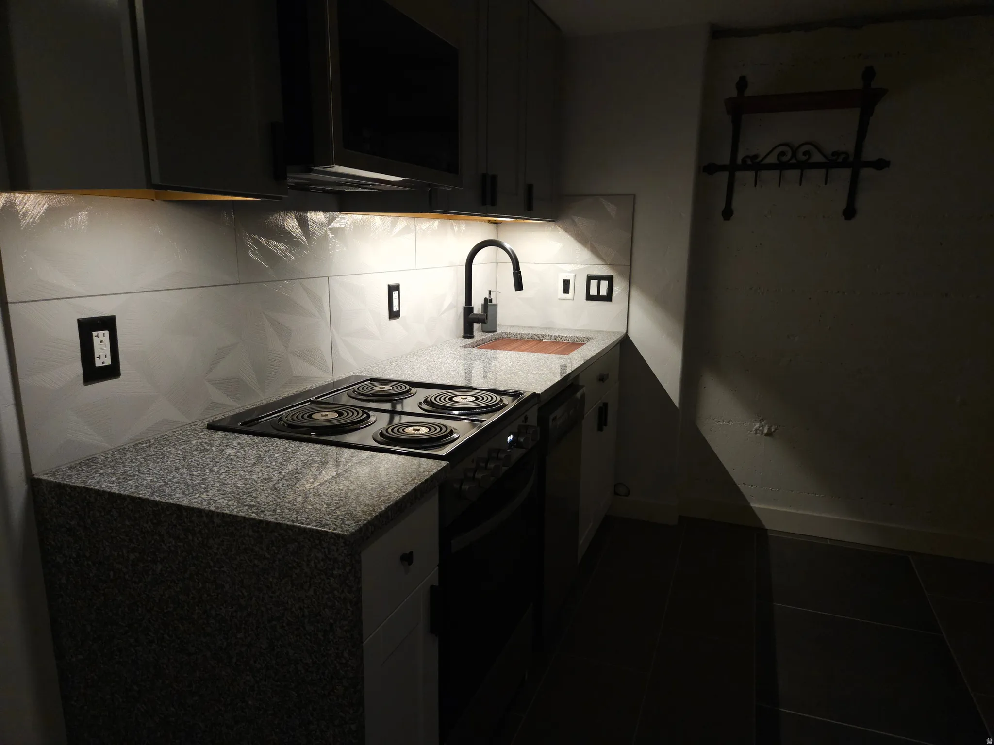 Compact kitchen featuring granite-look countertops, a four-burner electric range, and a geometric-patterned backsplash with under-cabinet lighting