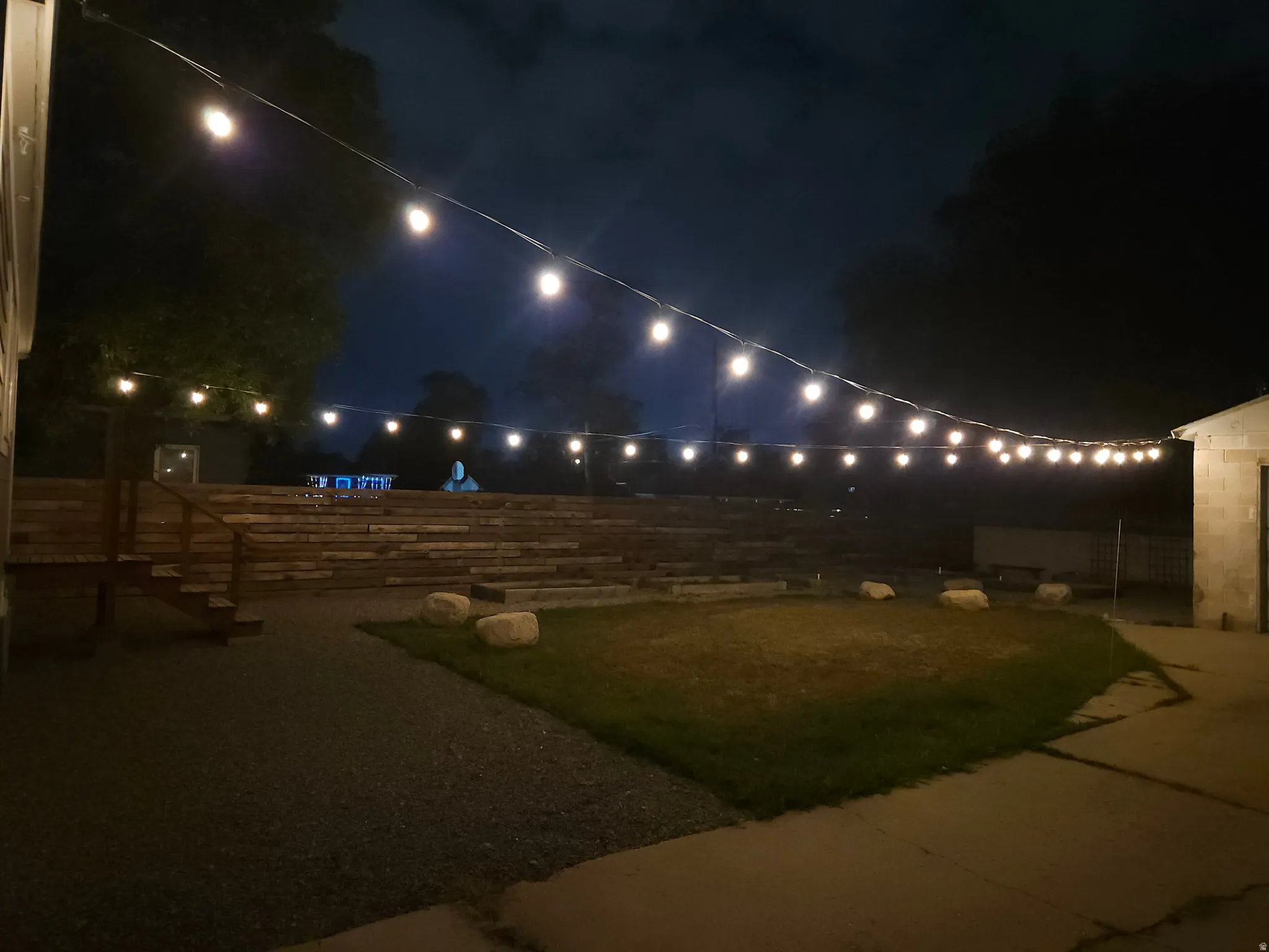 Outdoor area featuring string lighting, terraced wood landscaping, a gravel path, a concrete patio, and a grassy lawn