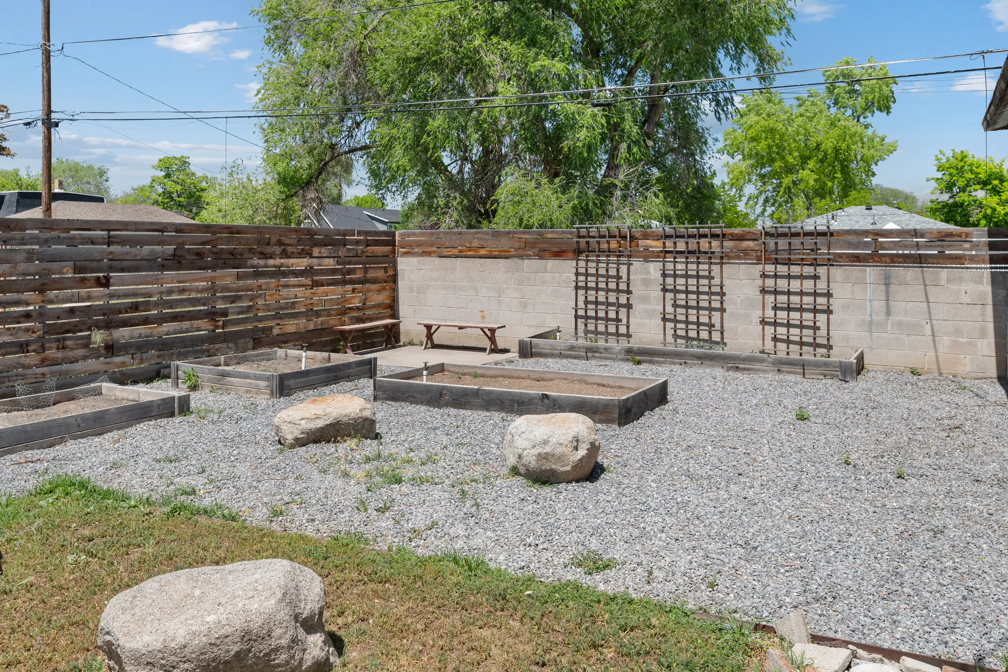 Gravel backyard featuring multiple raised garden beds, a horizontal slat wood fence, a concrete block wall with trellises, and natural stone accents